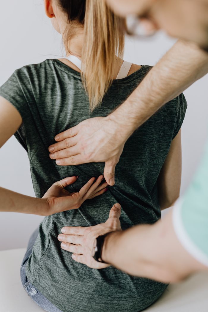 Home Top view of crop unrecognizable woman in casual wear showing chiropractor in eyeglasses and uniform pain spot on back while sitting on examination couch in hospital